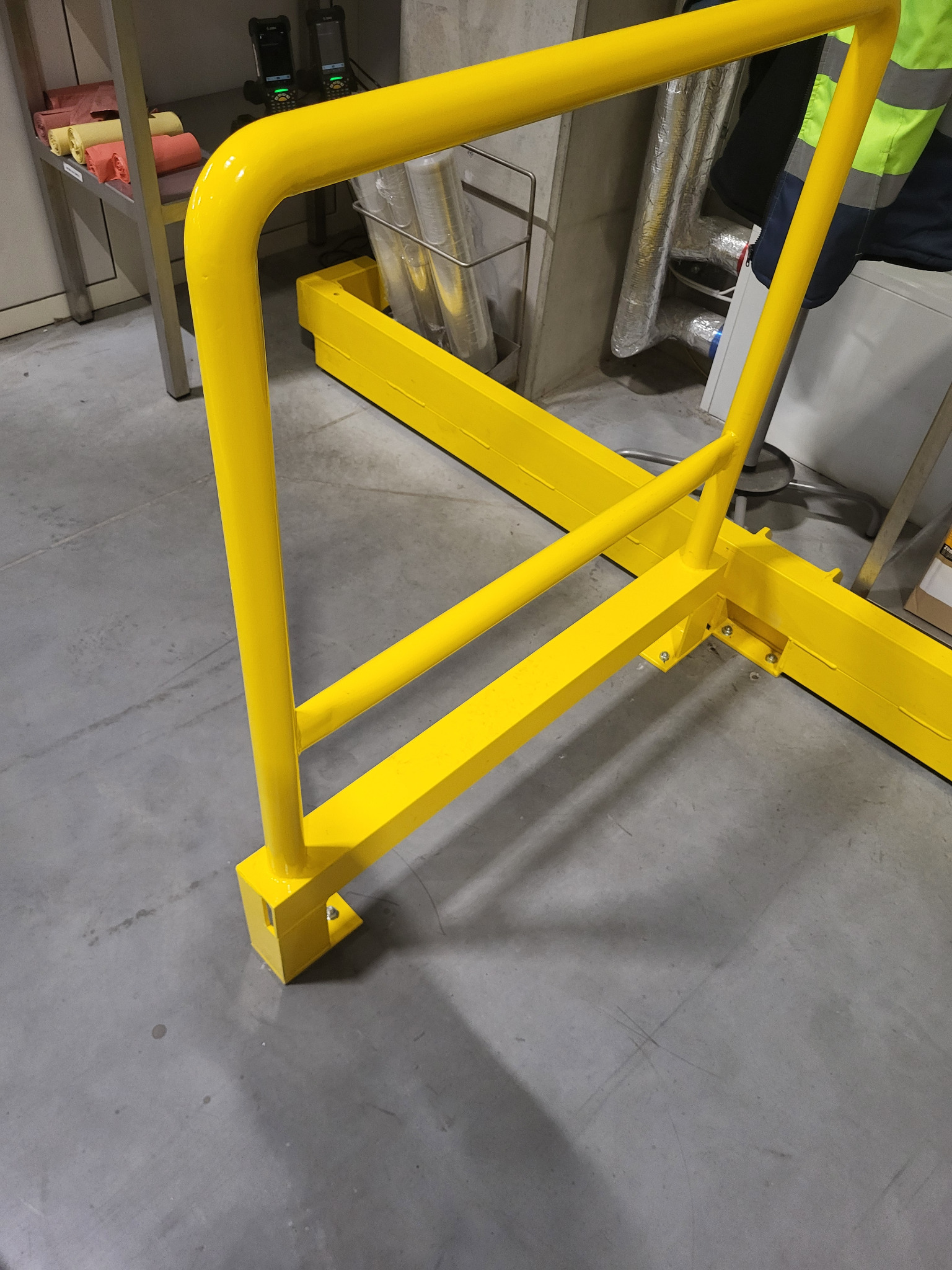 Żółta, metalowa barierka ochronna, zamontowana do szarej betonowej podłogi. Widoczne śruby mocujące. W tle instalacje i elementy wyposażenia.