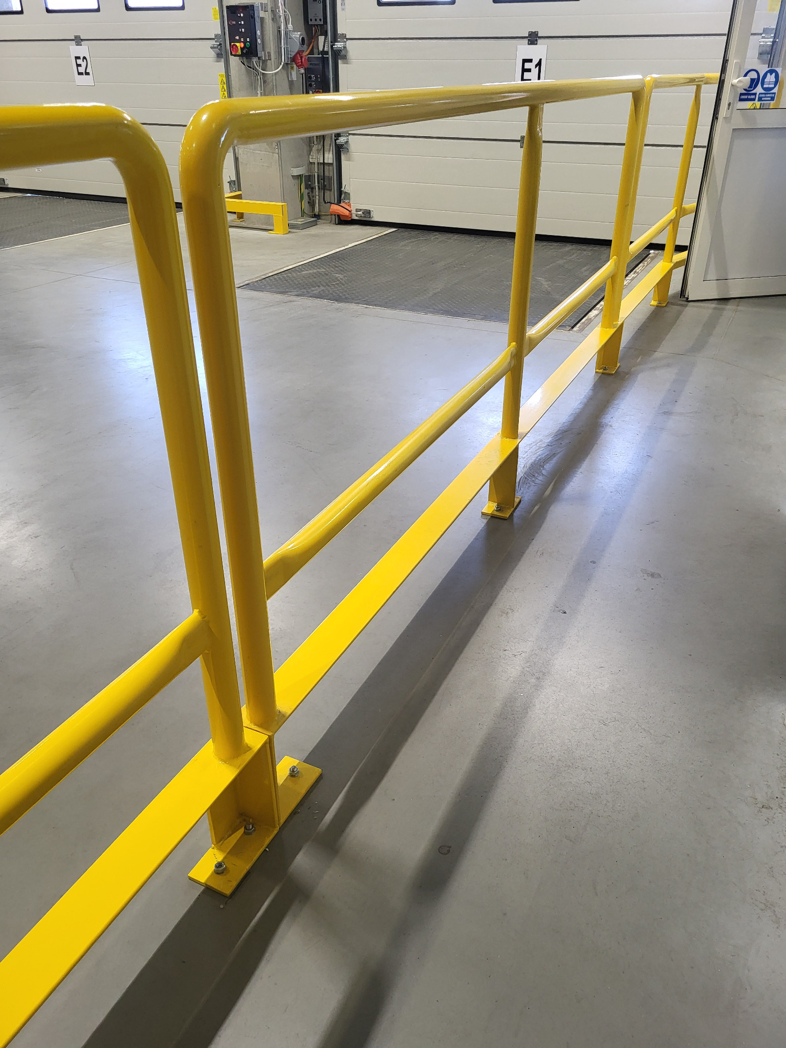 Żółta, metalowa balustrada ochronna w hali magazynowej, zamontowana przy bramie wjazdowej. Widoczne mocowania do podłoża i oznaczenia bram.