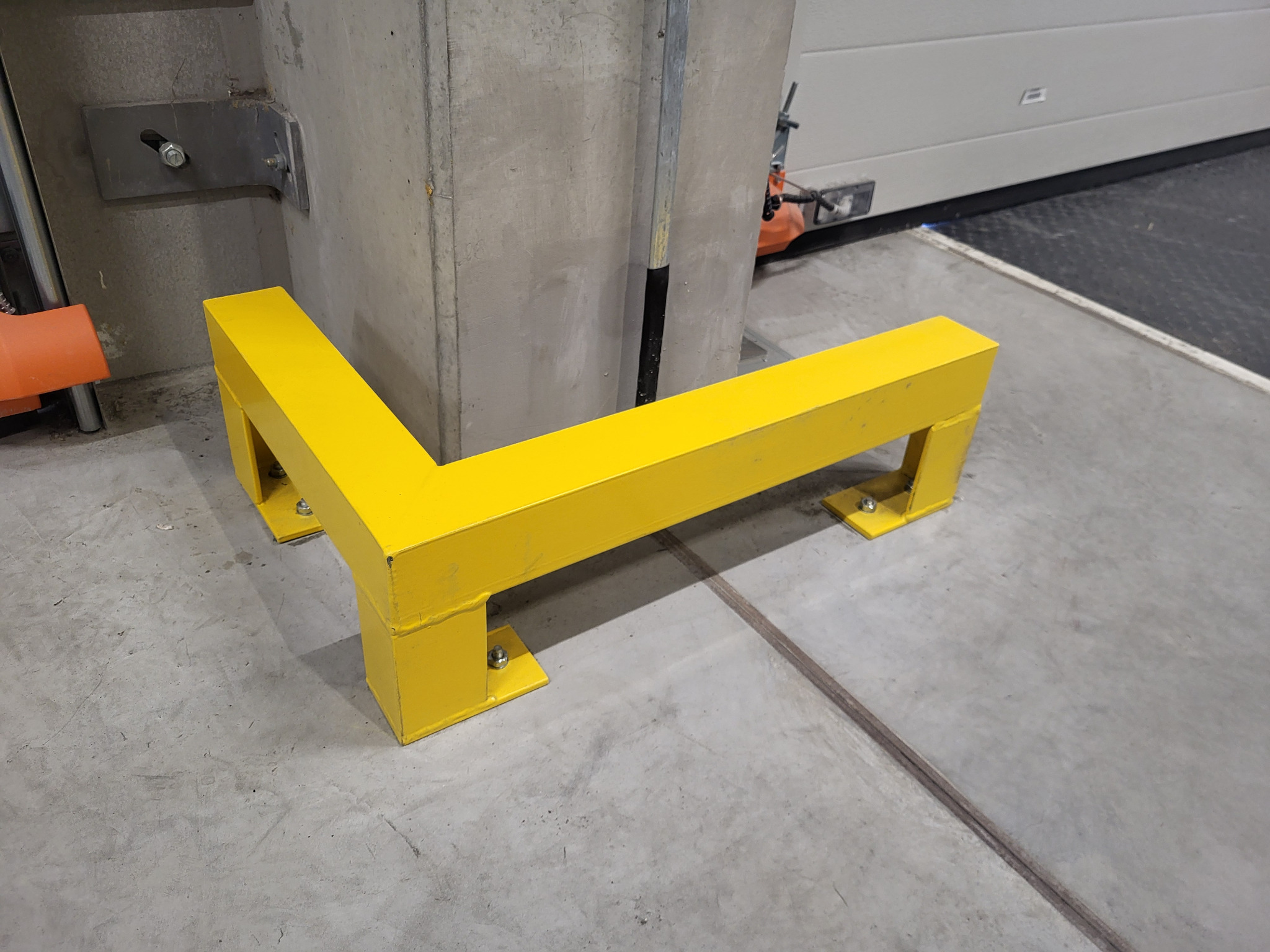 Żółta, spawana bariera ochronna z metalu, w kształcie litery L, zabezpieczająca betonowy słup w hali przemysłowej. Widoczne śruby mocujące do podłoża.