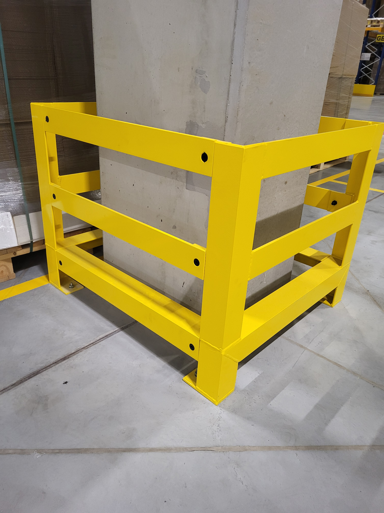 Żółta, stalowa osłona słupa w hali magazynowej. Trzy poziome belki tworzą ramę ochronną wokół betonowego słupa. Widoczne śruby mocujące. Tło stanowią regały i palety.