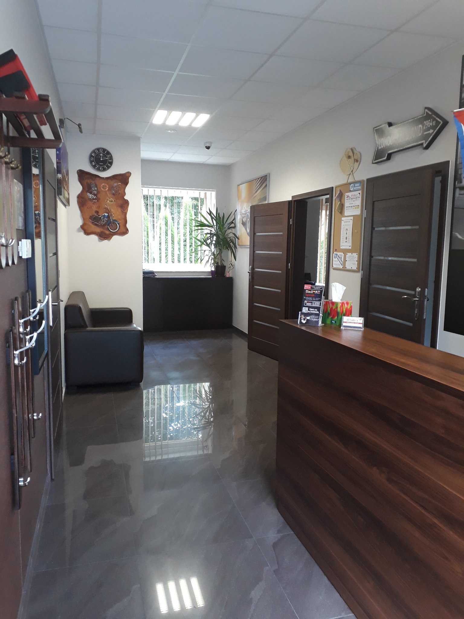 Recepcja w szkole językowej z ciemnymi meblami, na ścianie zegar i dekoracja z motocyklem, strzałka z napisem Londyn 2964 km.