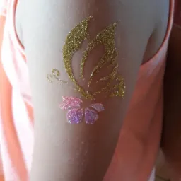 Złoty brokatowy motyl i różowy brokatowy kwiat na ramieniu dziecka, tymczasowy tatuaż.