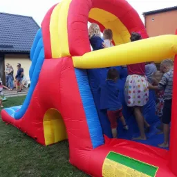 Dzieci w różnym wieku bawią się na dmuchanym zamku w kolorach czerwonym, żółtym i niebieskim, ustawionym na trawie przed budynkiem.