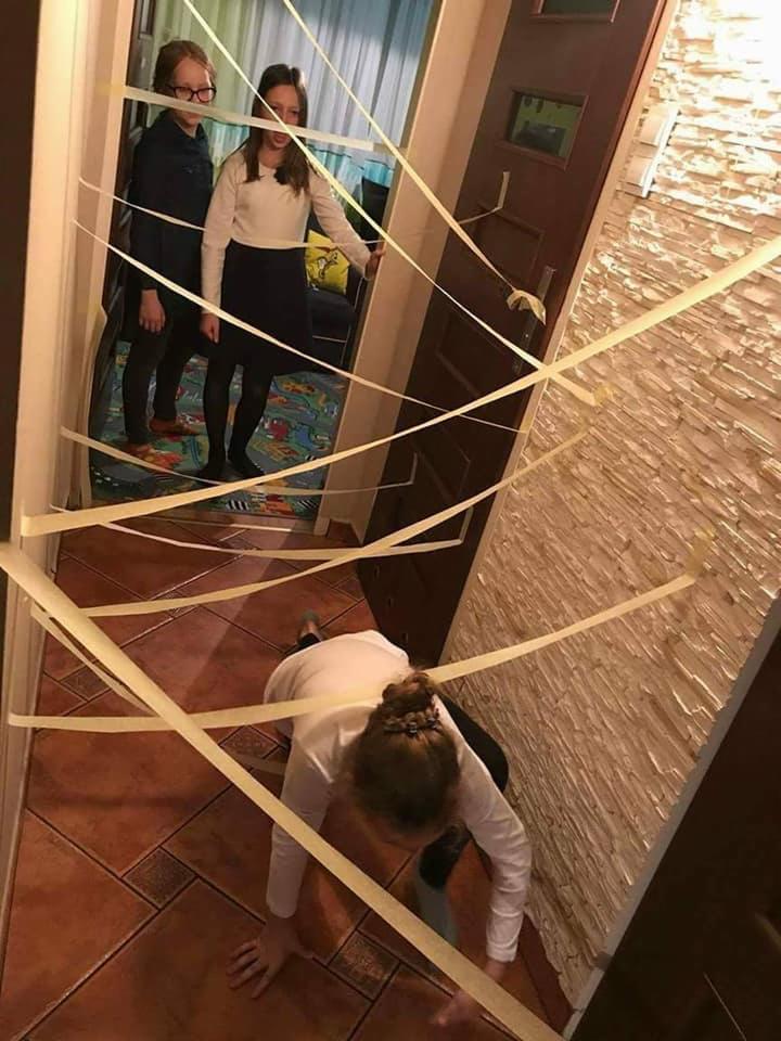 Dziewczyna czołga się pod taśmami w korytarzu, podczas gdy dwie inne dziewczyny stoją i obserwują, prawdopodobnie podczas zabawy lub gry.