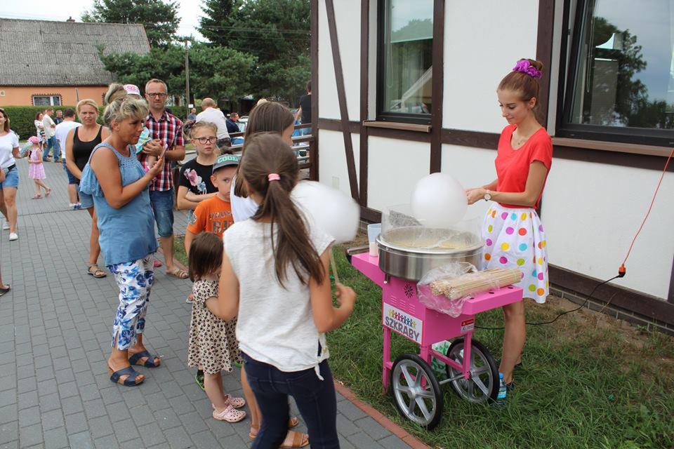 Dziewczyna w czerwonej koszulce i spódnicy w kropki przygotowuje watę cukrową na różowym wózku z napisem 'Szkraby' dla grupy dzieci i dorosłych.