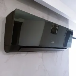 Czarny, błyszczący klimatyzator AUX zamontowany na ścianie o marmurkowym wzorze, widoczna ikona religijna odbijająca się w obudowie.