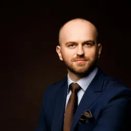 Portret mężczyzny w niebieskim garniturze, brązowym krawacie i poszetce, z krótką brodą i poważnym wyrazem twarzy na ciemnobrązowym tle.