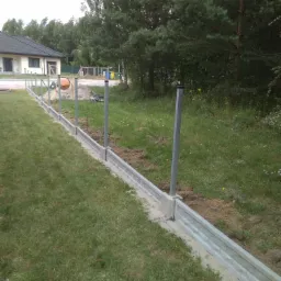 Wkopane prefabrykowane elementy betonowe pod ogrodzenie panelowe z metalowymi słupkami, widoczna betoniarka i przygotowany teren pod budowę ogrodzenia.