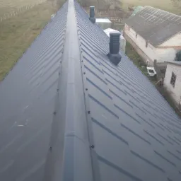 Widok z góry na ciemnoszary, metalowy dach z dwoma kominami wentylacyjnymi, na tle wiejskiego krajobrazu z budynkami gospodarczymi i polami.