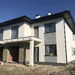 Dwukondygnacyjny dom bliźniak w trakcie prac elewacyjnych: część ściany pokryta styropianem z widocznymi kołkami, druga część pomalowana na biało z szarymi akcentami, okna zabezpieczone folią...