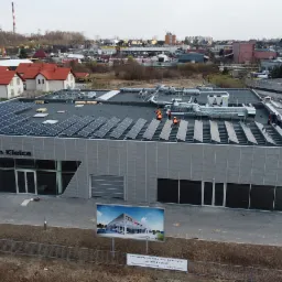 Montaż paneli fotowoltaicznych na dachu salonu Audi Centrum Kielce przez ekipę w pomarańczowych kamizelkach, widok z góry, w tle kominy przemysłowe i zabudowa miejska.