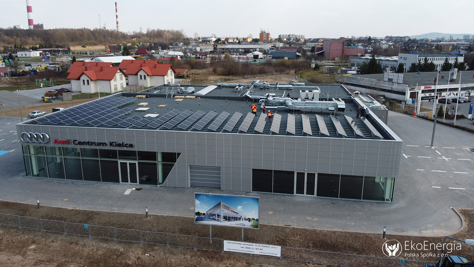 Montaż paneli fotowoltaicznych na dachu salonu Audi Centrum Kielce przez ekipę w pomarańczowych kamizelkach, widok z góry, w tle kominy przemysłowe i zabudowa miejska.