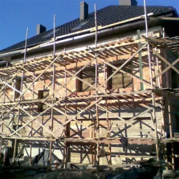 Wznoszenie dwukondygnacyjnego budynku z czerwonej cegły, z widocznym rusztowaniem i fragmentem dachu z ciemnej dachówki oraz kominami w słoneczny dzień.