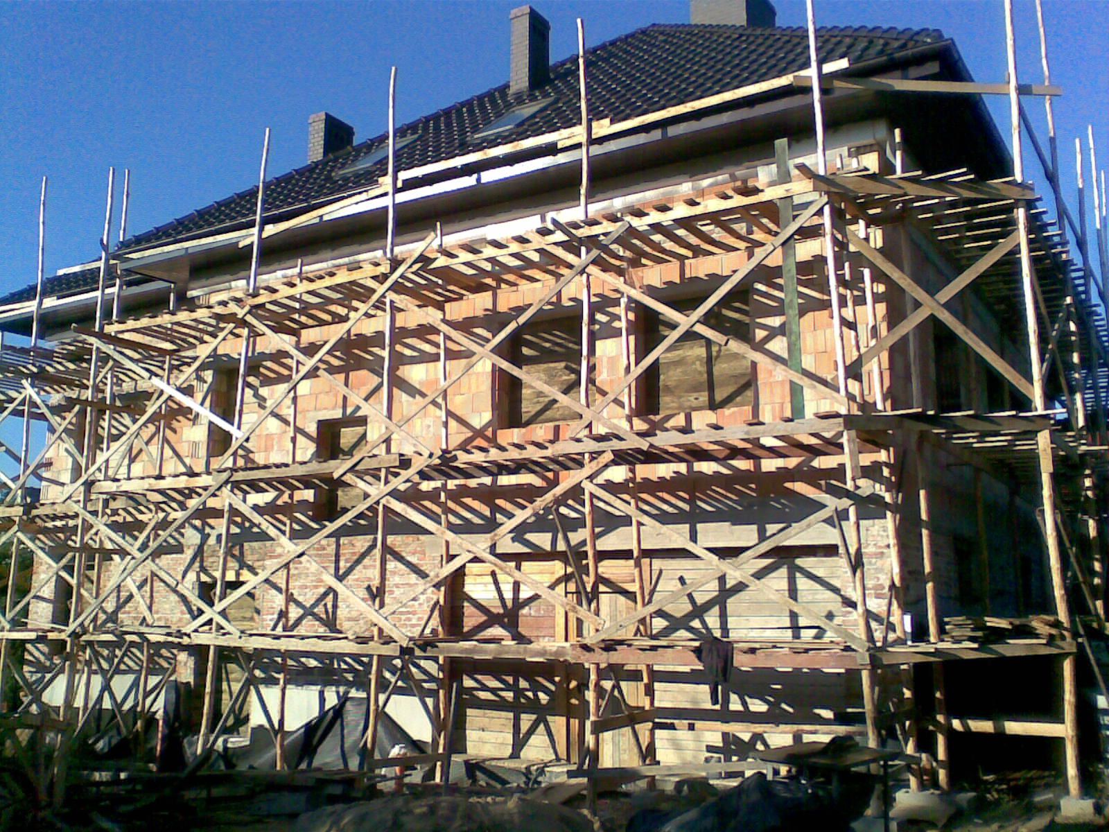 Wznoszenie dwukondygnacyjnego budynku z czerwonej cegły, z widocznym rusztowaniem i fragmentem dachu z ciemnej dachówki oraz kominami w słoneczny dzień.