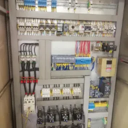 Rozbudowana szafa sterownicza z licznymi przekaźnikami, bezpiecznikami i przewodami w kolorach niebieskim, żółtym i czerwonym, zamontowana w szarej obudowie.