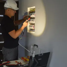 Mężczyzna w kasku i okularach ochronnych, mierzy napięcie w skrzynce rozdzielczej za pomocą miernika, na stole obok laptop i narzędzia.