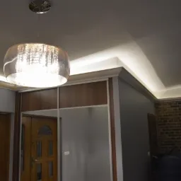 Kryształowy żyrandol wiszący pod sufitem z oświetleniem LED, widoczne drzwi wejściowe i ściana z cegły