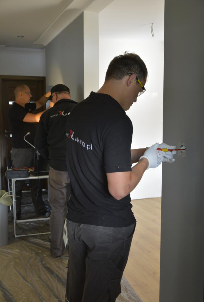 Elektryk w okularach ochronnych, w firmowej koszulce z logo, montuje gniazdko elektryczne w szarej ścianie, używając izolowanego śrubokrętu; w tle widoczni dwaj inni elektrycy podczas pracy.