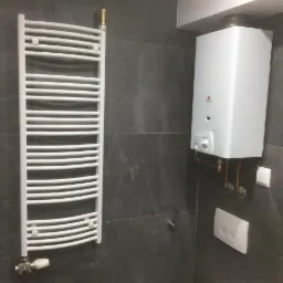 Biały grzejnik drabinkowy i wiszący kocioł gazowy na szarej, płytkowej ścianie w łazience, widoczne miedziane rury instalacji.