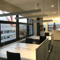 Jasne biuro z rzędami białych biurek i czarnymi krzesłami, widok na port z pomarańczowym dźwigiem przez duże okna.