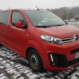Czerwony Citroen SpaceTourer na kostce brukowej z resztkami śniegu, widok z przodu na tle drzew i pochmurnego nieba.