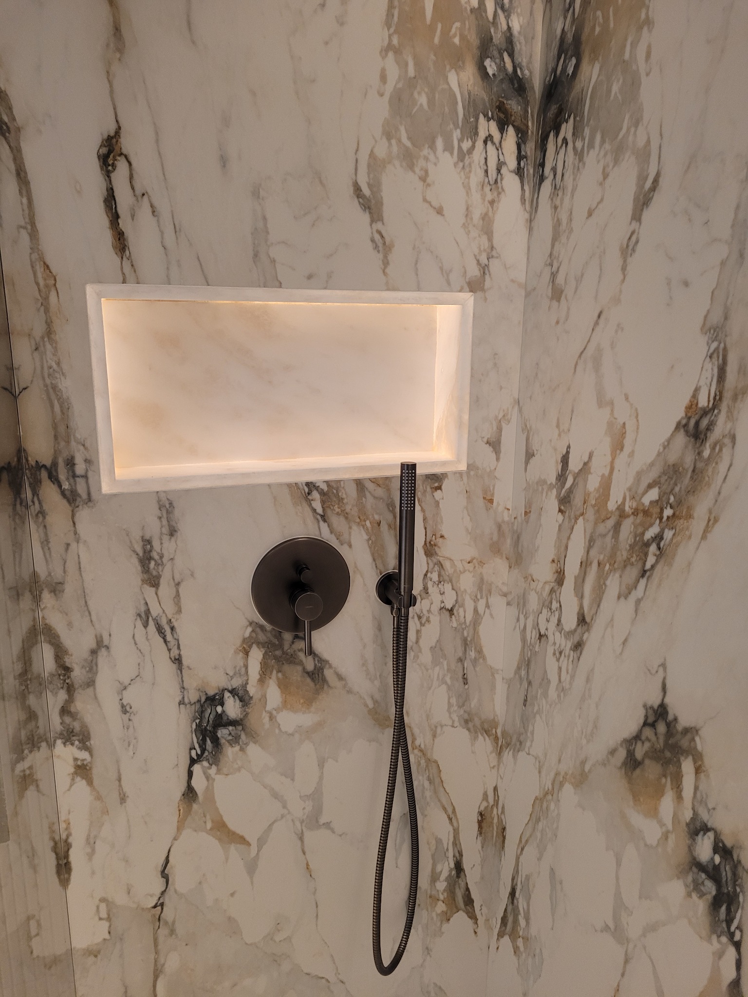 Wnęka z podświetleniem w marmurowej ścianie kabiny prysznicowej, ciemna bateria i słuchawka prysznicowa.