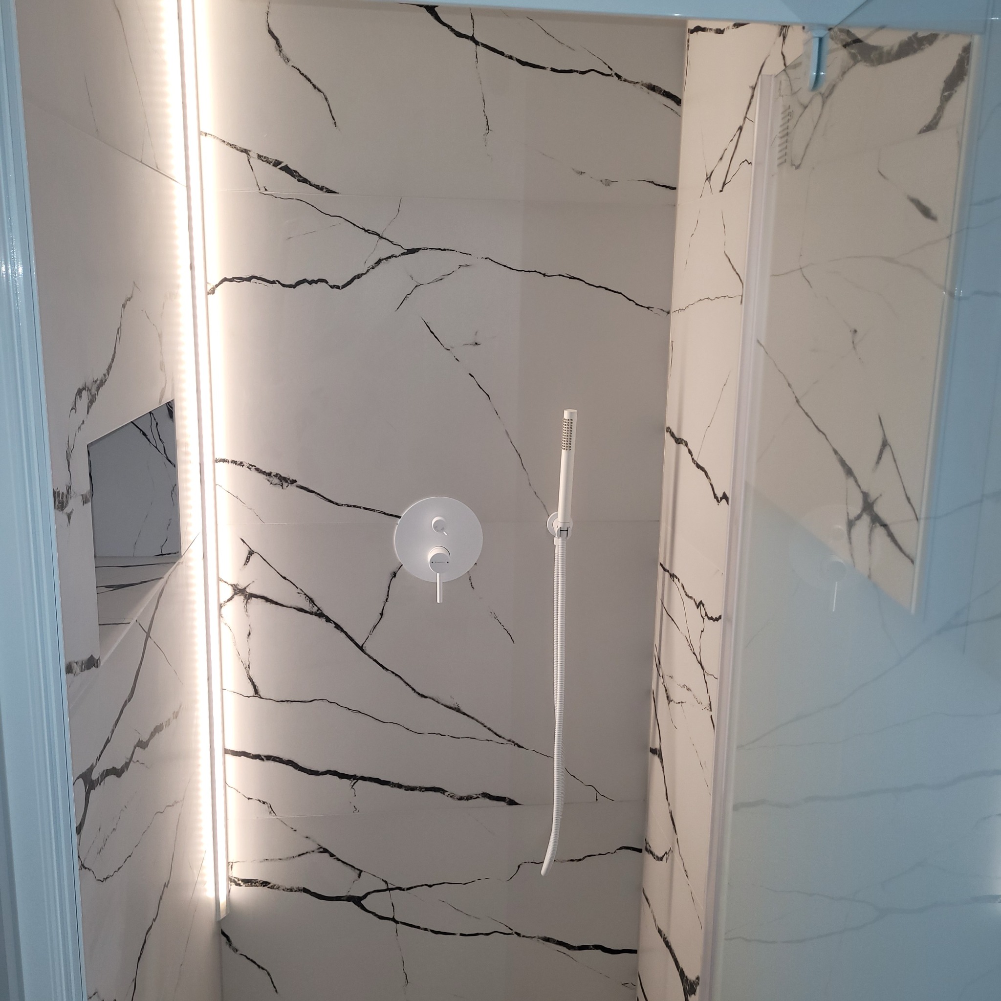 Kabina prysznicowa z białymi płytkami imitującymi marmur z czarnymi żyłami, minimalistyczna bateria podtynkowa i deszczownica, oświetlenie LED w narożniku.