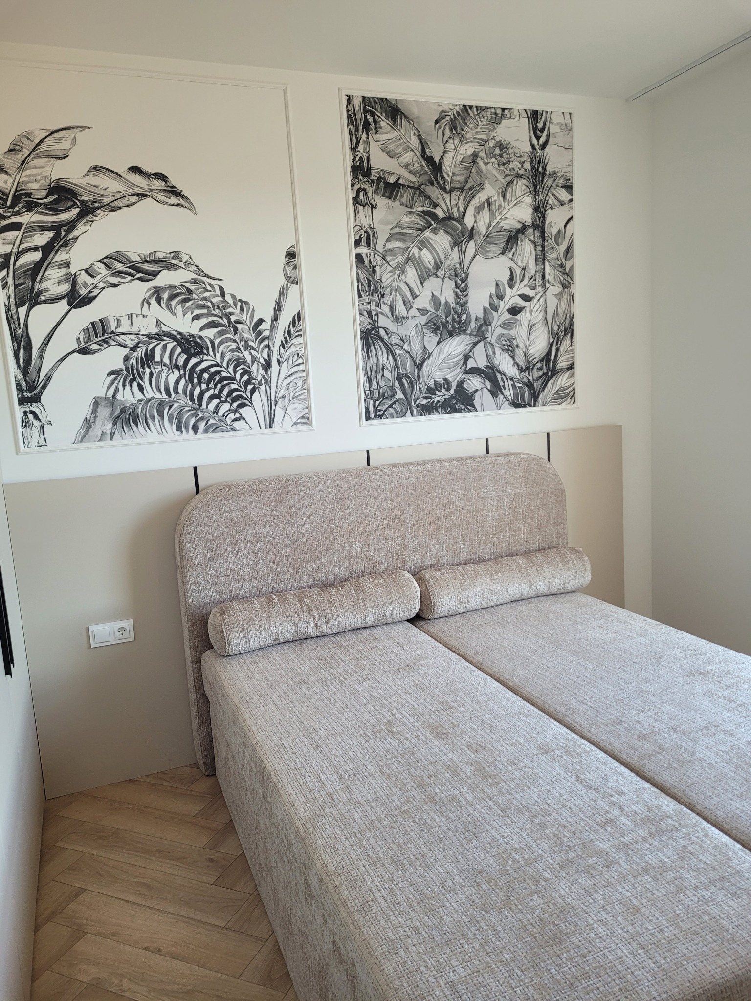 Jasna sypialnia z dwoma łóżkami pojedynczymi, wezgłowie w kolorze beżowym, na ścianie grafiki z motywem roślinnym, parkiet ułożony w jodełkę.