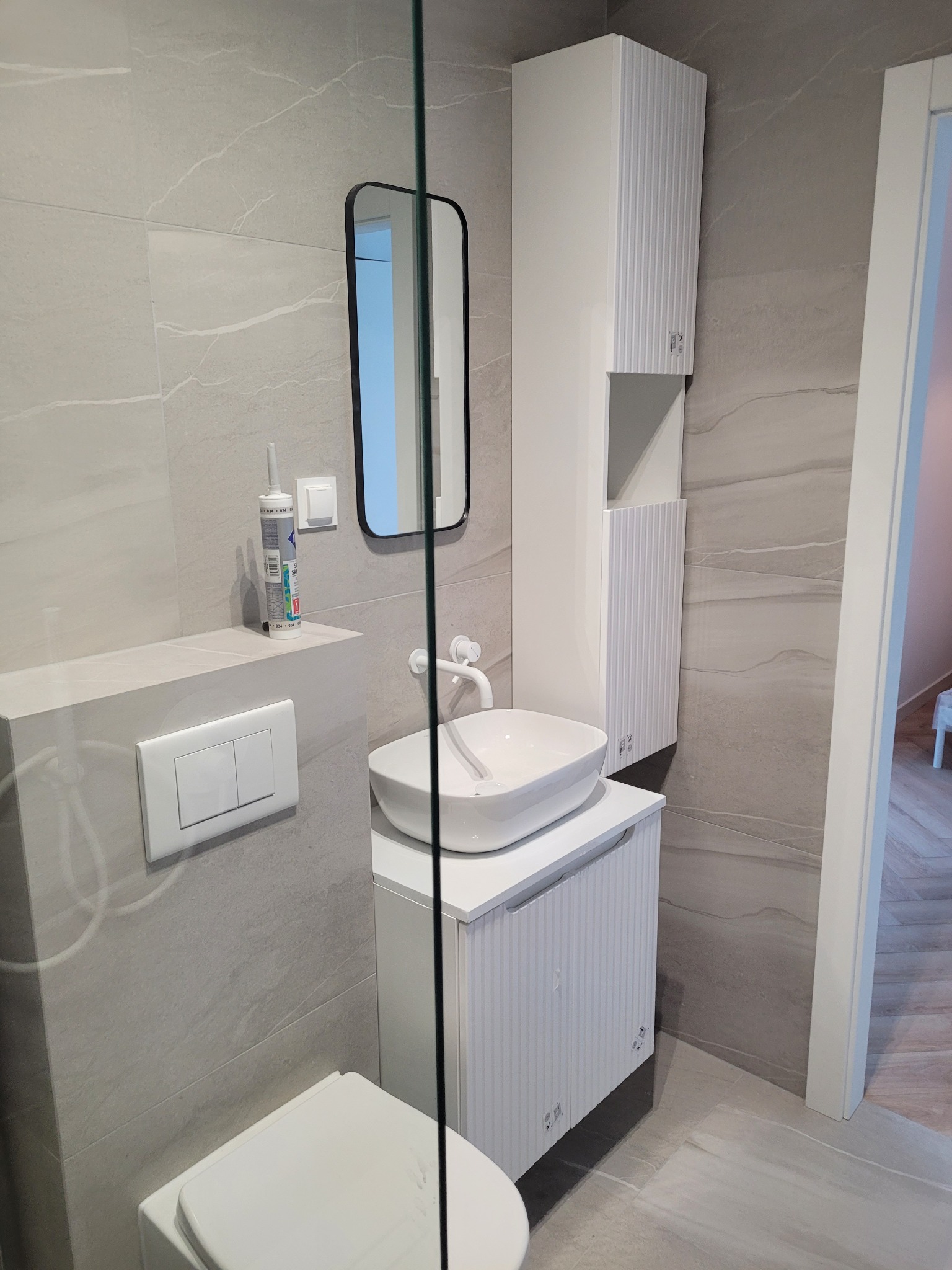 Nowoczesna łazienka w odcieniach szarości z białą armaturą, wiszącą szafką z umywalką nablatową, wysoką szafką oraz czarną ramą lustra, silikon sanitarny na półce, widoczne płytki imitujące marmur...