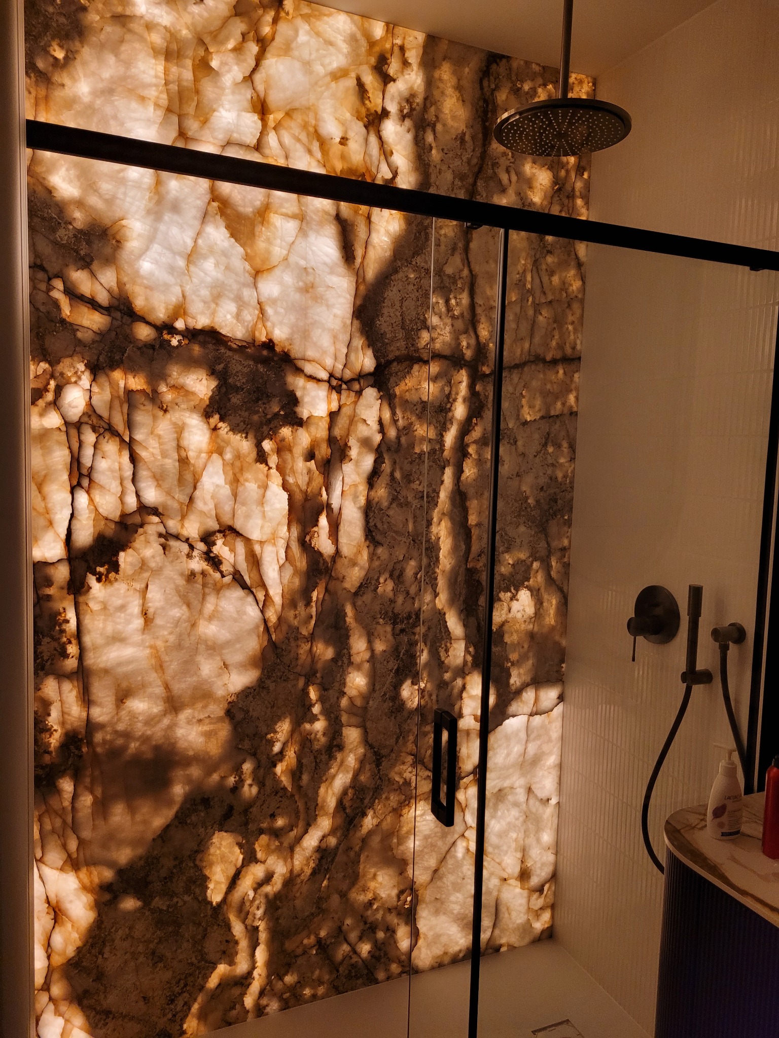 Podświetlana ściana z onyksu w kabinie prysznicowej z czarną armaturą i szklanymi drzwiami.