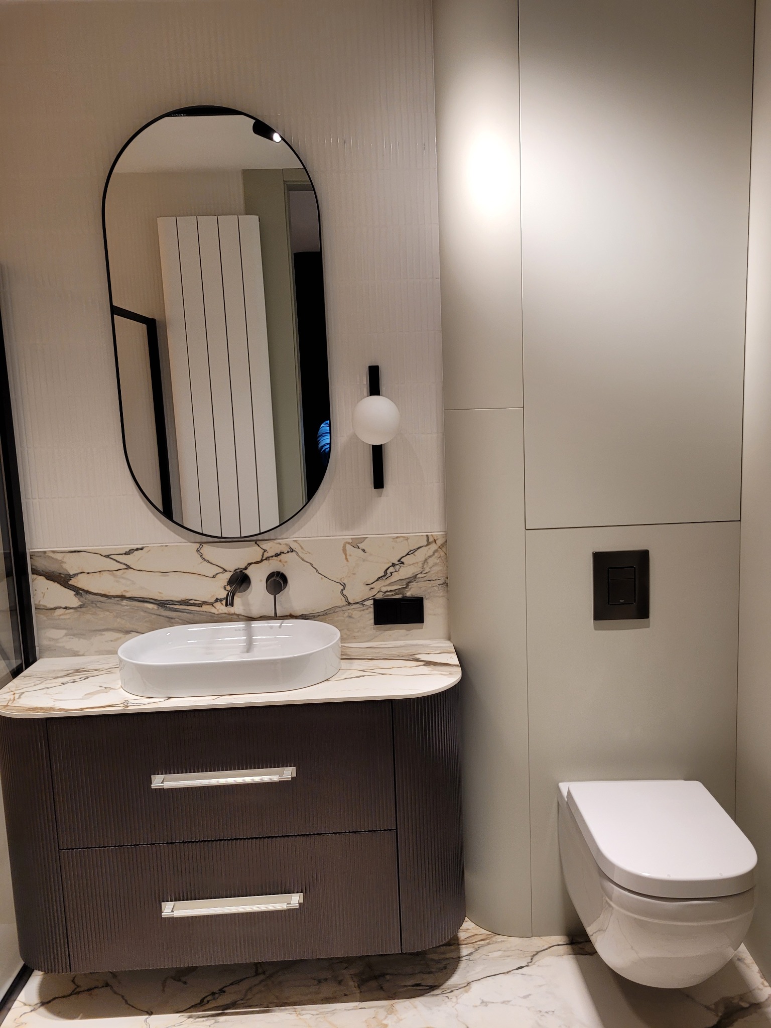 Nowoczesna łazienka z umywalką nablatową, owalnym lustrem w czarnej ramie, toaletą podwieszaną i szafką z ryflowanym frontem w odcieniach brązu, wykończona płytkami imitującymi marmur.