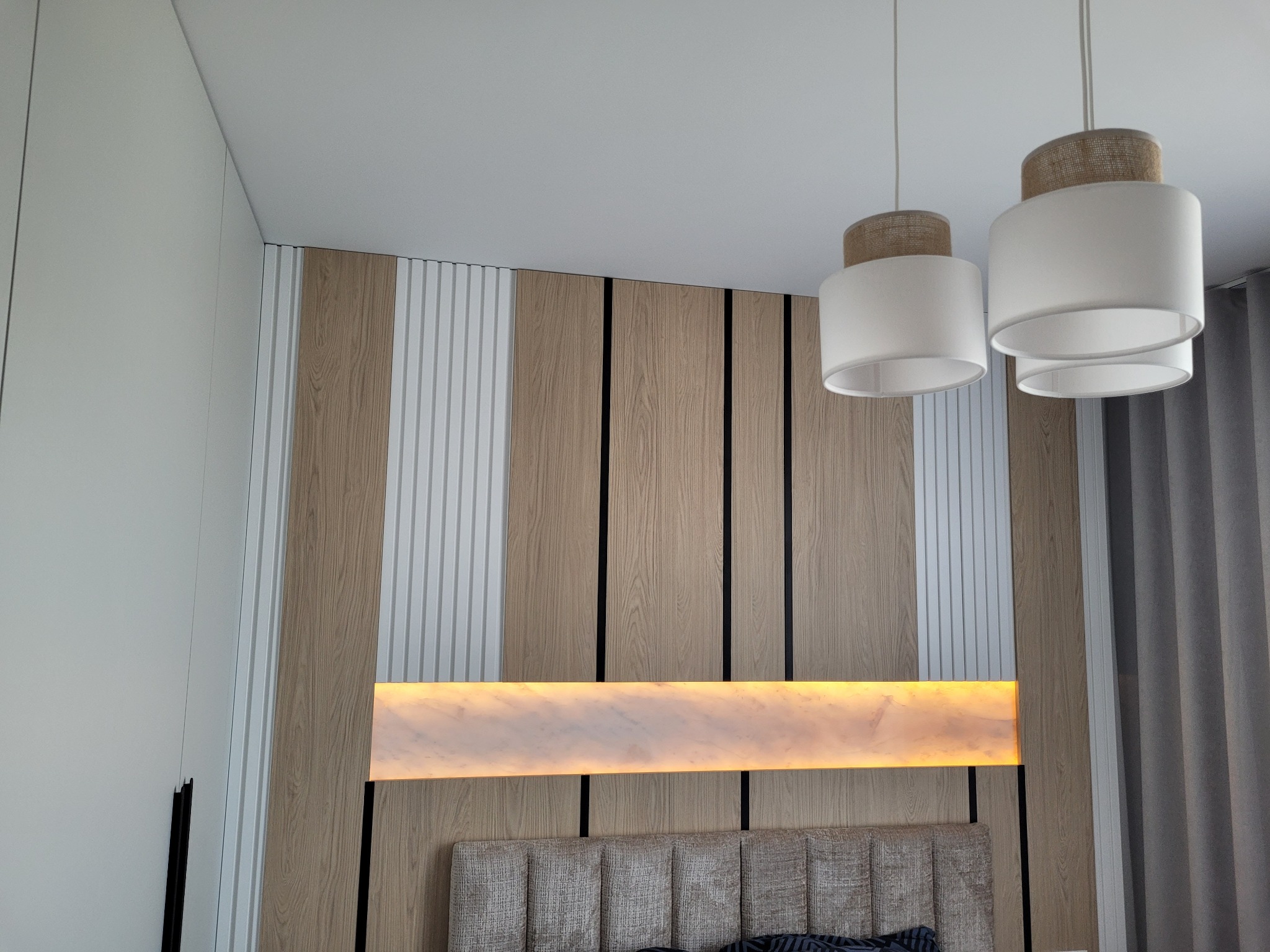 Nowoczesna sypialnia: dekoracyjna ściana z paneli drewnianych i białych, oświetlenie LED, lampy wiszące i zasłony. Widok na zagłówek łóżka.