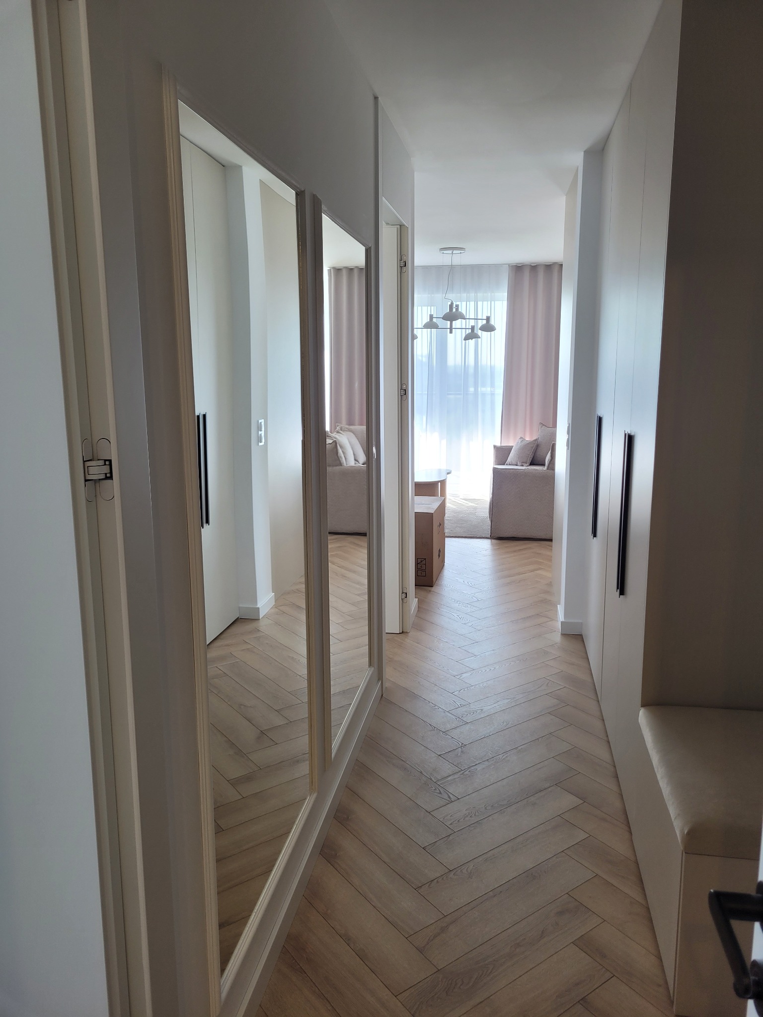 Jasny korytarz z parkietem w jodełkę, lustrami i szafami w zabudowie. W tle widoczny salon z sofą i zasłonami. Minimalistyczny design, neutralna kolorystyka.