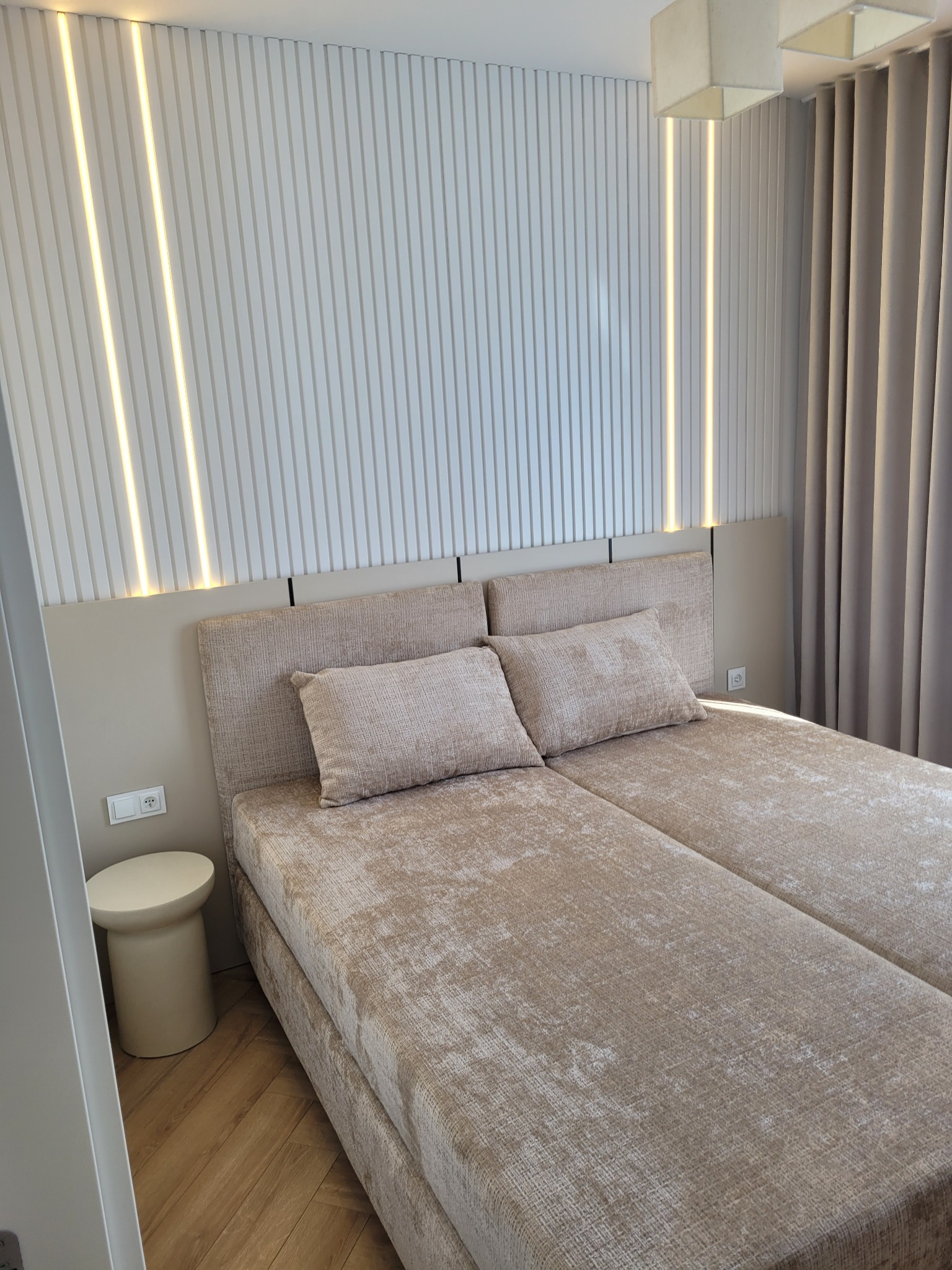 Nowoczesna sypialnia: beżowe łóżko z zagłówkiem, ryflowana ściana z oświetleniem LED, drewniana podłoga i zasłony w tonacji beżowej. Minimalistyczny stolik nocny.