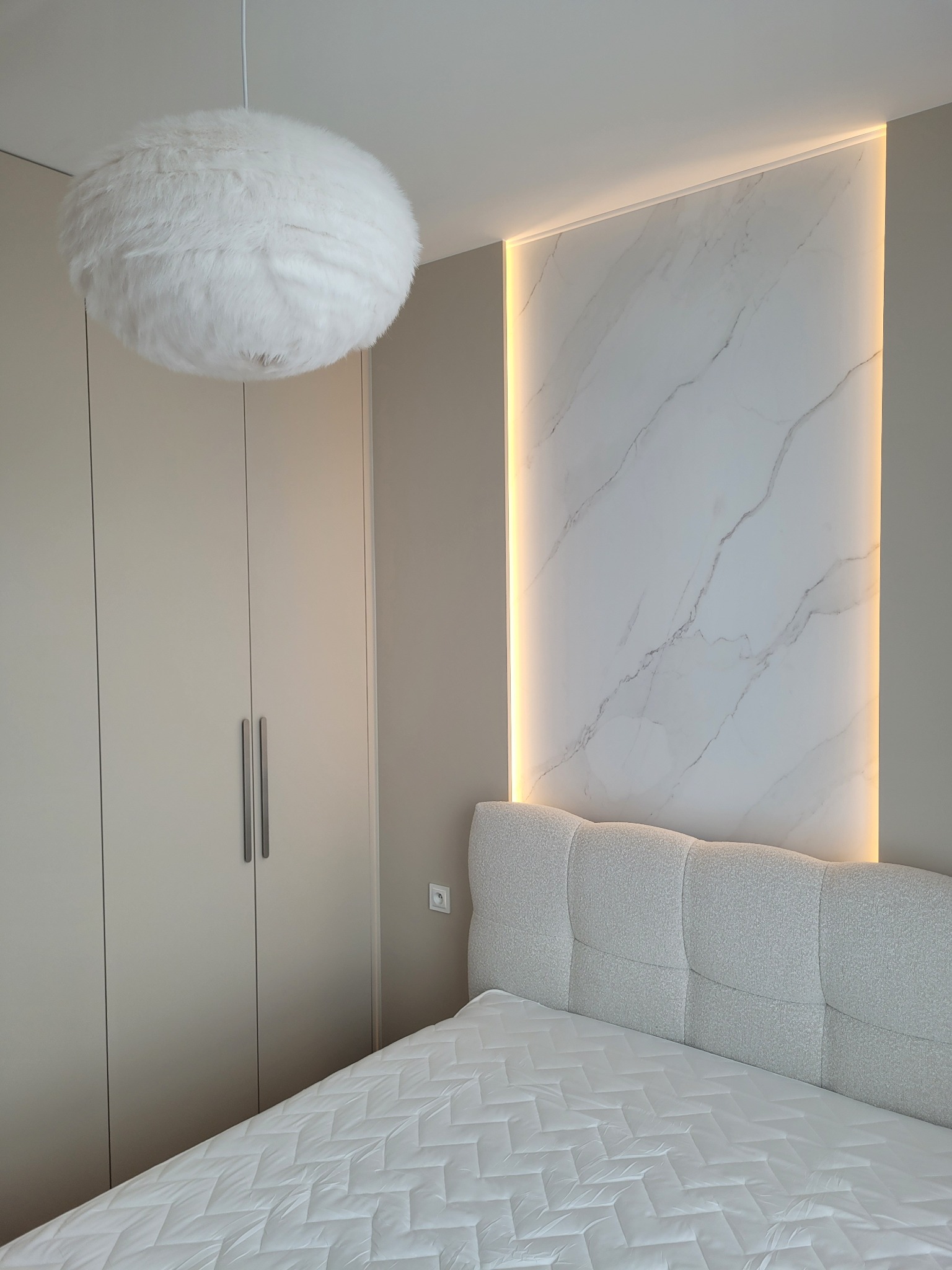 Sypialnia z beżową szafą, łóżkiem z tapicerowanym zagłówkiem i marmurową ścianą z podświetleniem LED. Nad łóżkiem puszysta, okrągła lampa.