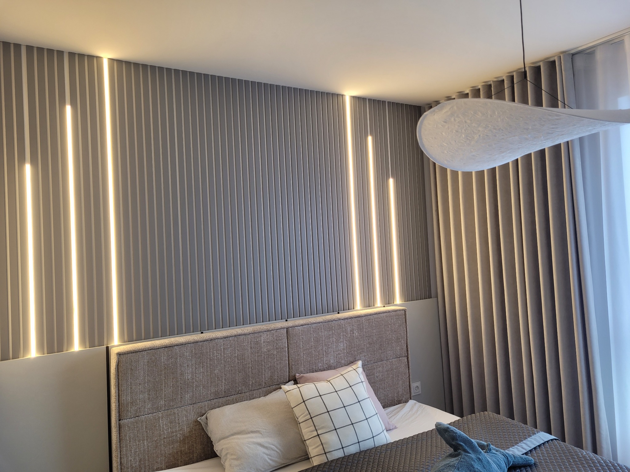 Nowoczesna sypialnia: szare panele ścienne z oświetleniem LED, designerska lampa, zasłony w odcieniach szarości, tapicerowane wezgłowie łóżka i dekoracyjne poduszki.