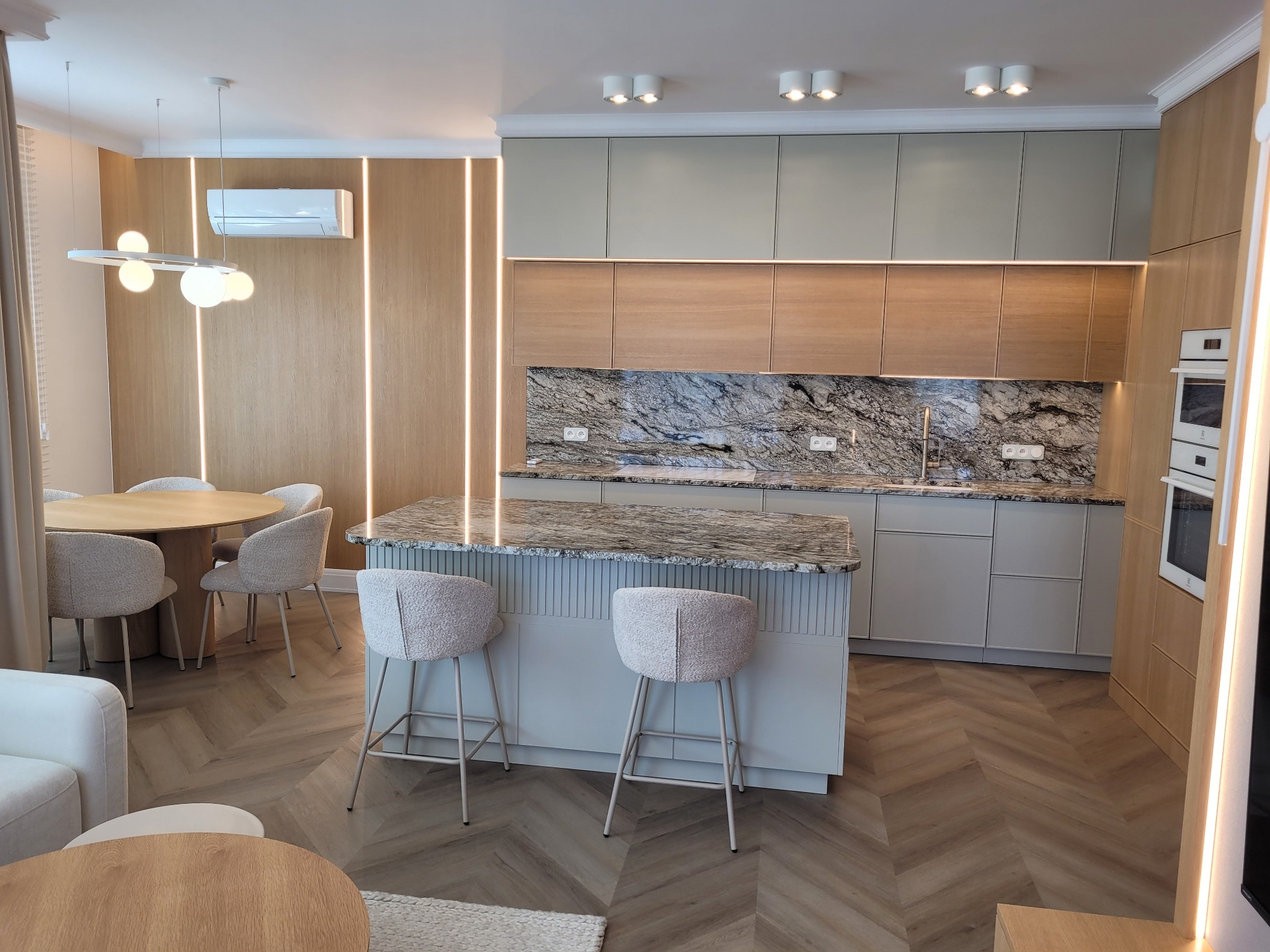 Nowoczesne wnętrze: jadalnia z okrągłym stołem i wyspą kuchenną z marmurowym blatem, parkiet w jodełkę, oświetlenie LED i klimatyzacja.