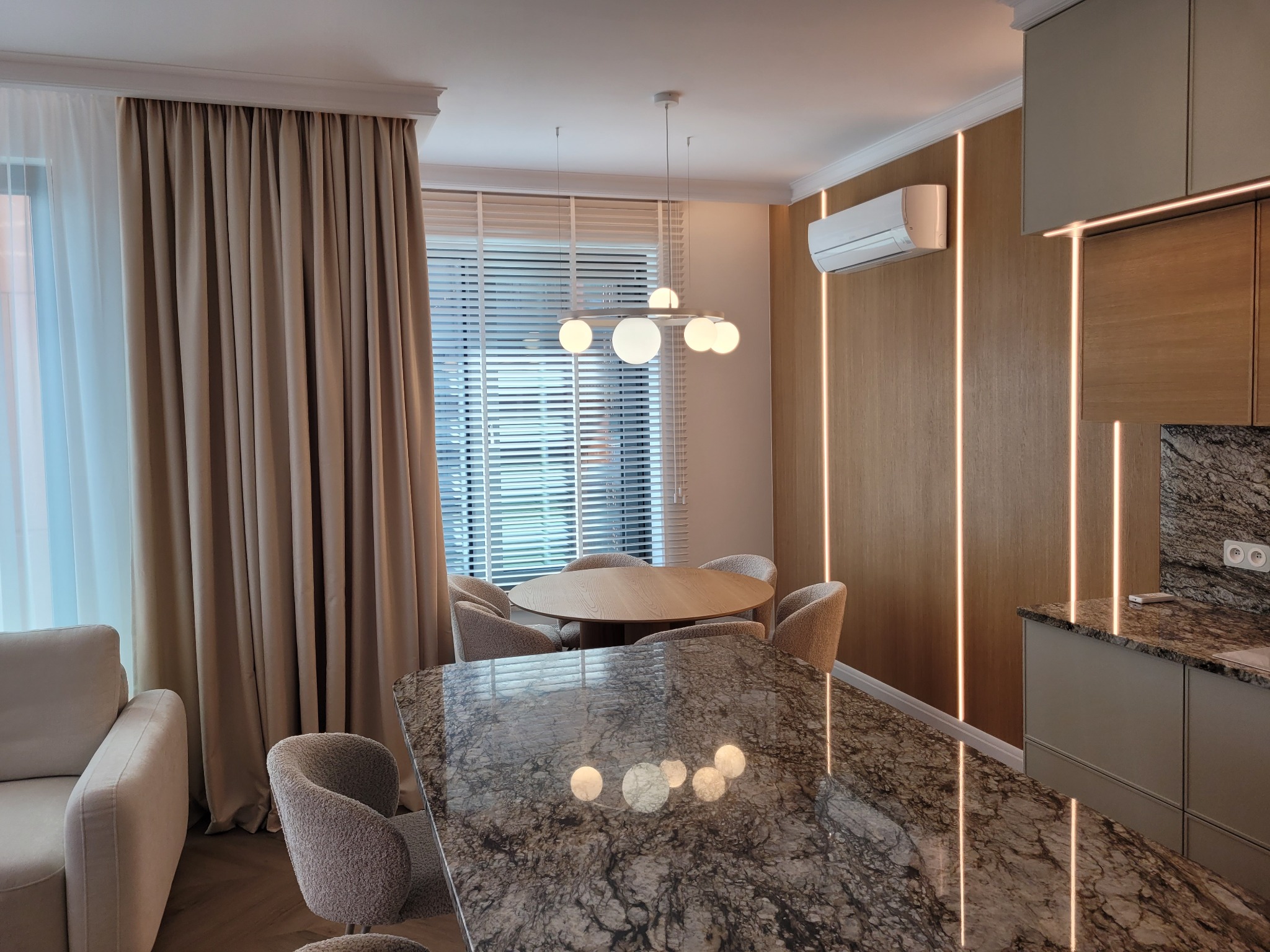 Eleganckie wnętrze jadalni z blatem z granitu, oświetleniem LED w pionowych pasach na ścianie i okrągłym stołem z krzesłami w neutralnych barwach. Nowoczesny design.