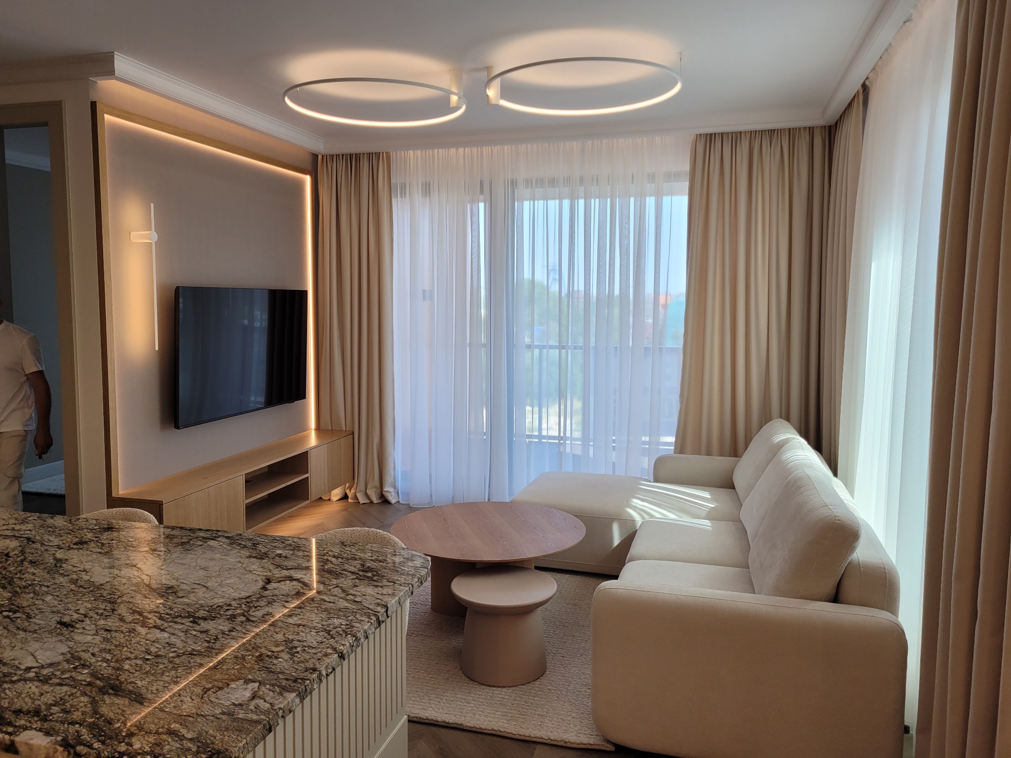 Jasny salon z beżową sofą, okrągłym stolikiem kawowym i telewizorem podświetlonym LED. Widoczne zasłony i lampy sufitowe w kształcie okręgów. Minimalistyczny design.