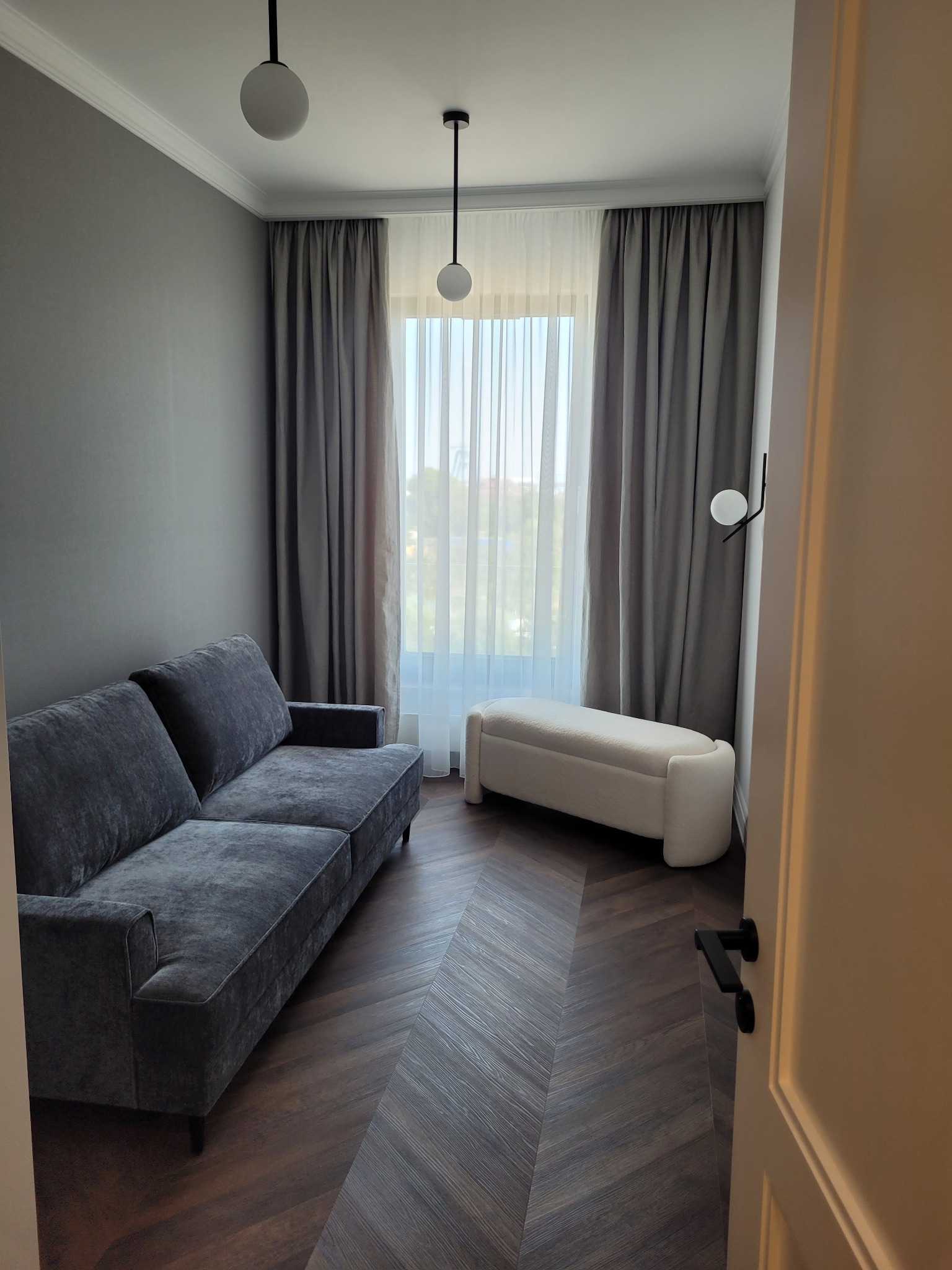 Stylowy salon z szarą sofą, parkietem w jodełkę i białymi zasłonami. Minimalistyczne lampy sufitowe i kinkiet dodają elegancji. Widok zza otwartych drzwi.