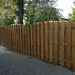 Długi, drewniany płot z zaokrąglonymi deskami, zabezpieczony bejcą w odcieniach brązu, oddzielający posesję od chodnika z kostki brukowej, z widocznym drzewem w tle.