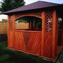 ALBERO - Drewniana altana ogrodowa z ażurowymi ścianami i ciemnym dachem na tle trawnika i zabudowań.