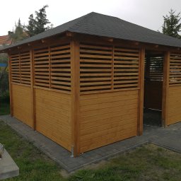 ALBERO - Drewniana altana ogrodowa z czarnym, spadzistym dachem z gontu bitumicznego, częściowo osłonięta poziomymi listwami, posadowiona na szarej kostce brukowej, z widocznym fragmentem ogrodu w tle.