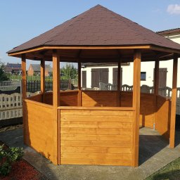 ALBERO - Ośmiokątna, drewniana altana z brązowym, bitumicznym dachem, częściowo obudowana deskami, stojąca na betonowej wylewce w ogrodzie.