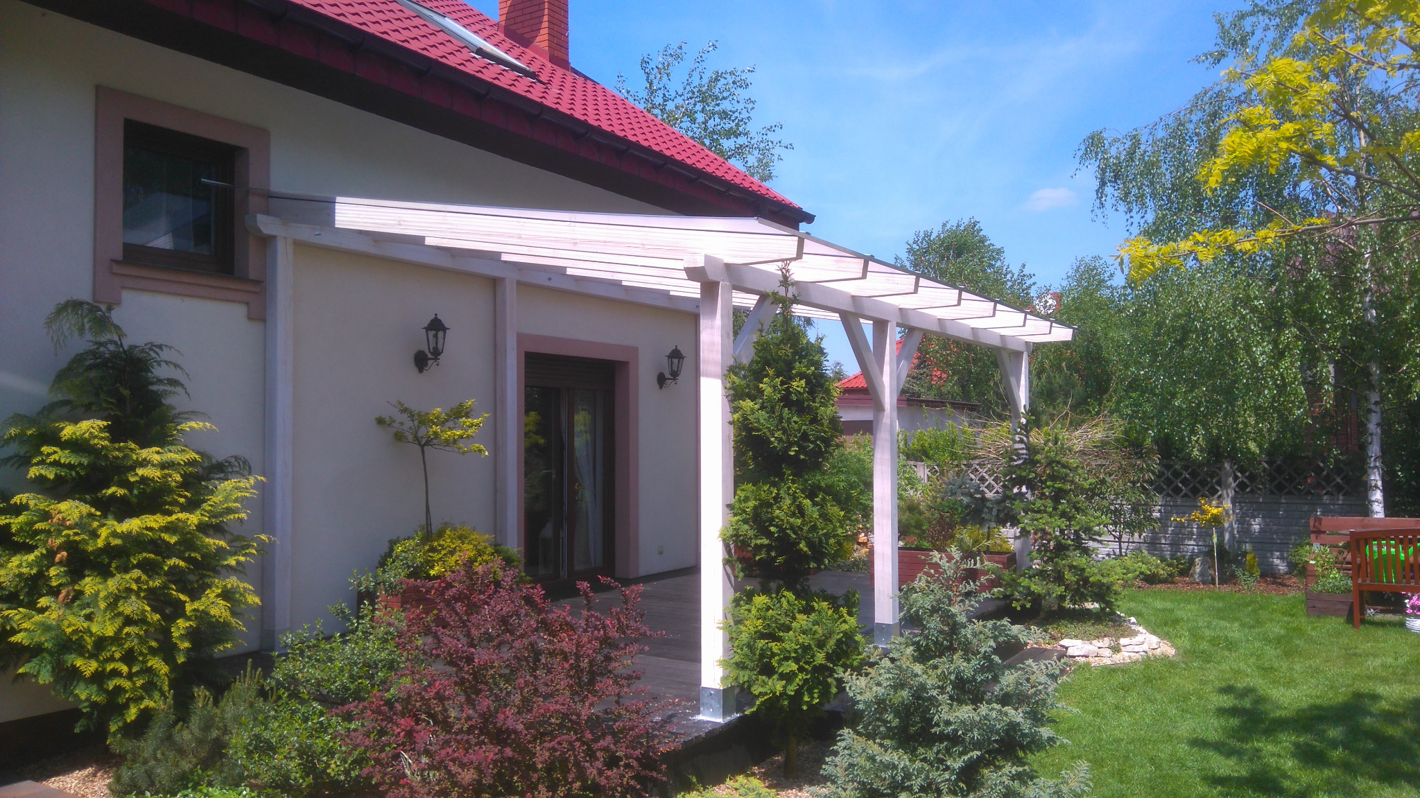 Biała, drewniana pergola przy domu z czerwonym dachem, otoczona zadbanym ogrodem z różnorodną roślinnością i trawnikiem.