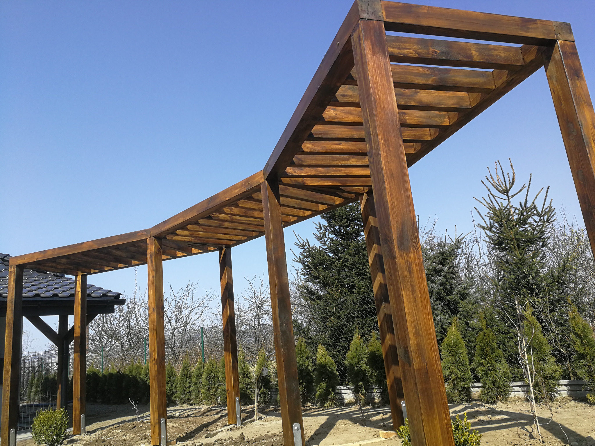 Drewniana konstrukcja pergoli z widocznym zadaszeniem altany w tle, zamontowana na metalowych kotwach w gruncie, na tle błękitnego nieba i zieleni ogrodu.