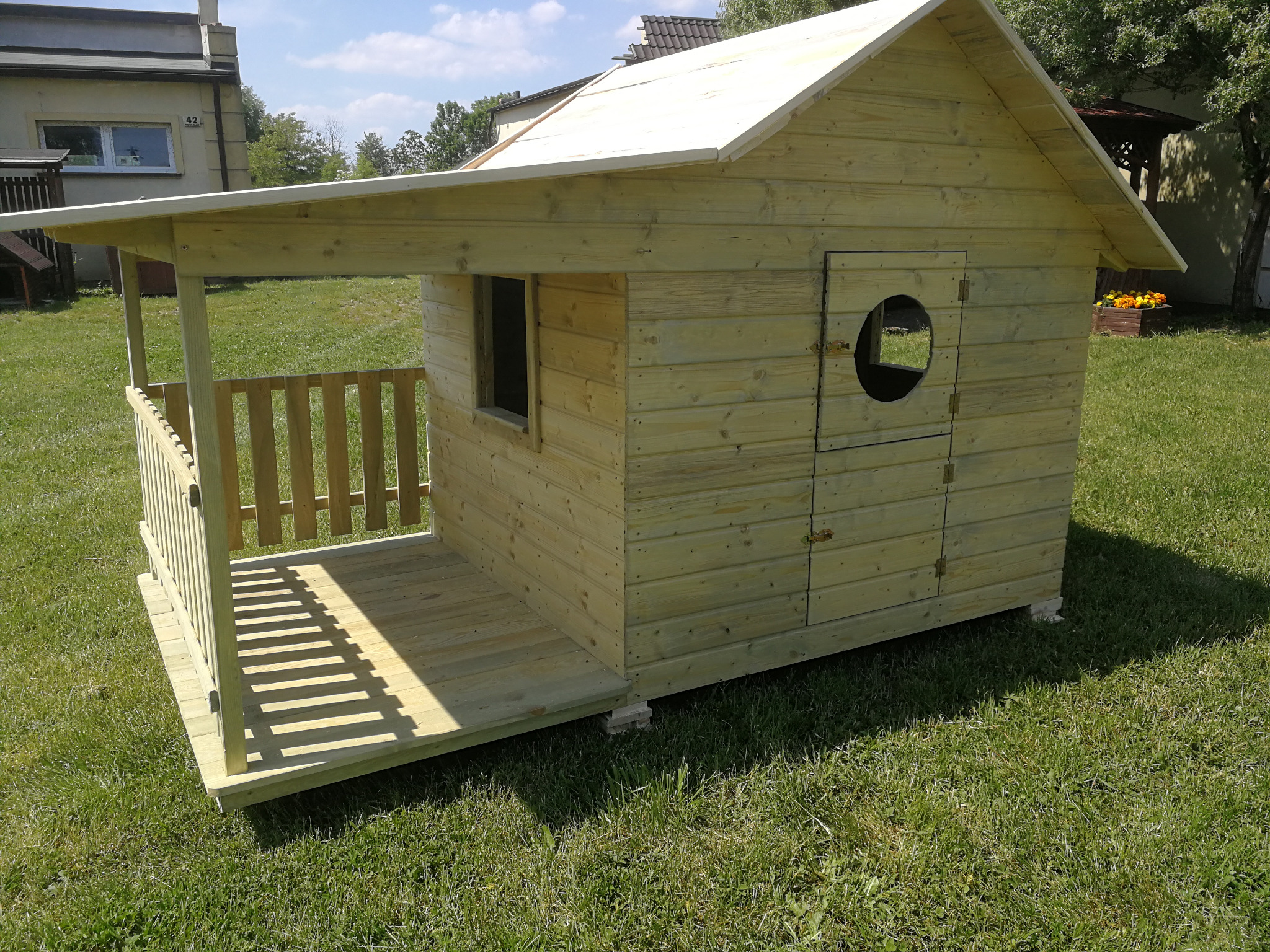 Drewniany domek dla dzieci z małym gankiem, okrągłym okienkiem i dwuspadowym dachem, ustawiony na trawniku przed budynkiem mieszkalnym.