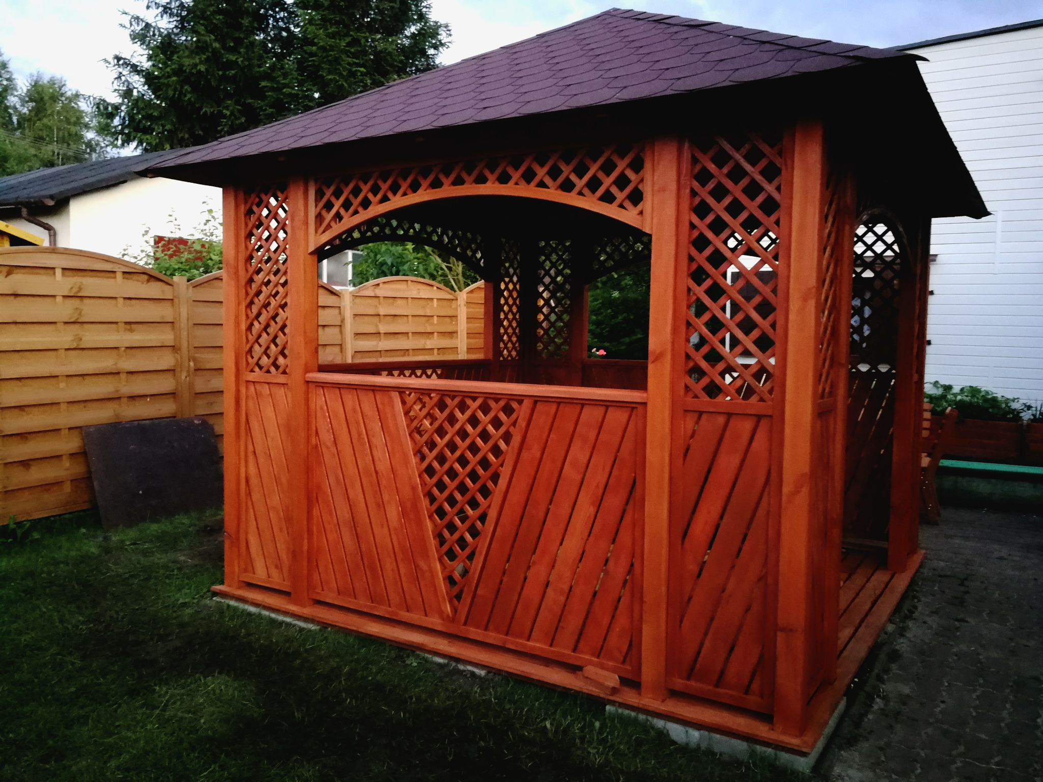 Drewniana altana ogrodowa z ażurowymi ścianami i ciemnym dachem na tle trawnika i zabudowań.