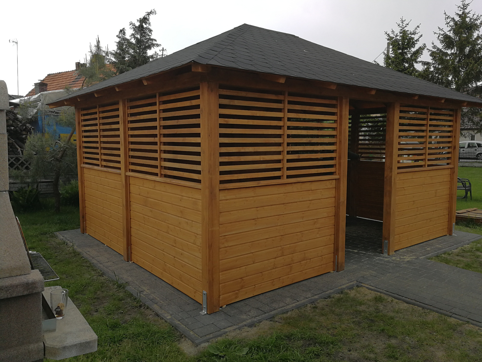 Drewniana altana ogrodowa z czarnym, spadzistym dachem z gontu bitumicznego, częściowo osłonięta poziomymi listwami, posadowiona na szarej kostce brukowej, z widocznym fragmentem ogrodu w tle.