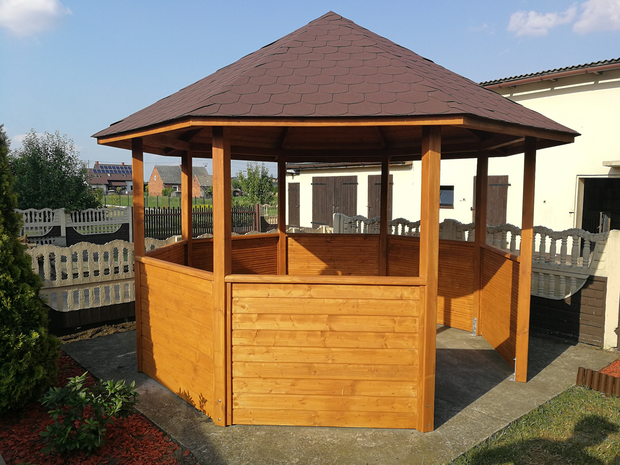 Ośmiokątna, drewniana altana z brązowym, bitumicznym dachem, częściowo obudowana deskami, stojąca na betonowej wylewce w ogrodzie.